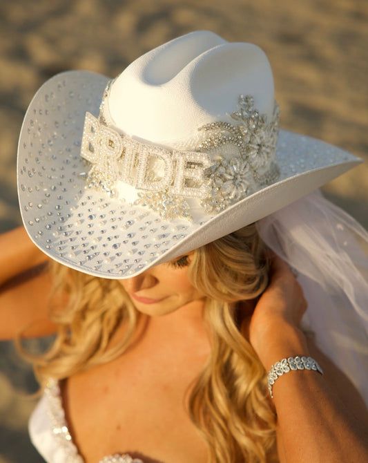 a woman wearing a white cowboy hat on the beach with her wedding dress blowing in the wind