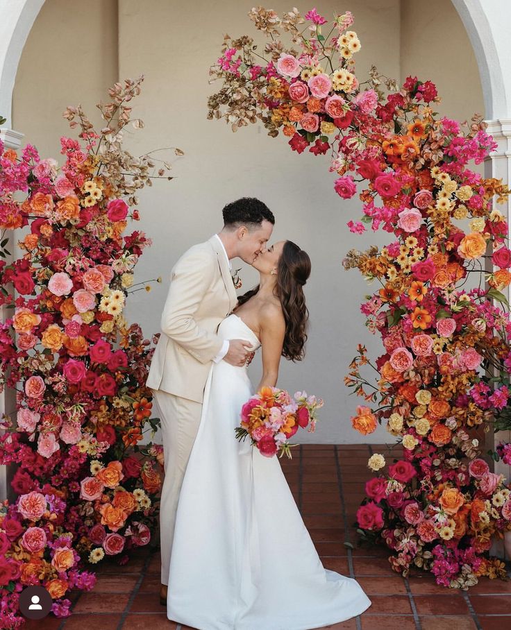 a bride and groom kissing in front of an archway with flowers on the wall behind them