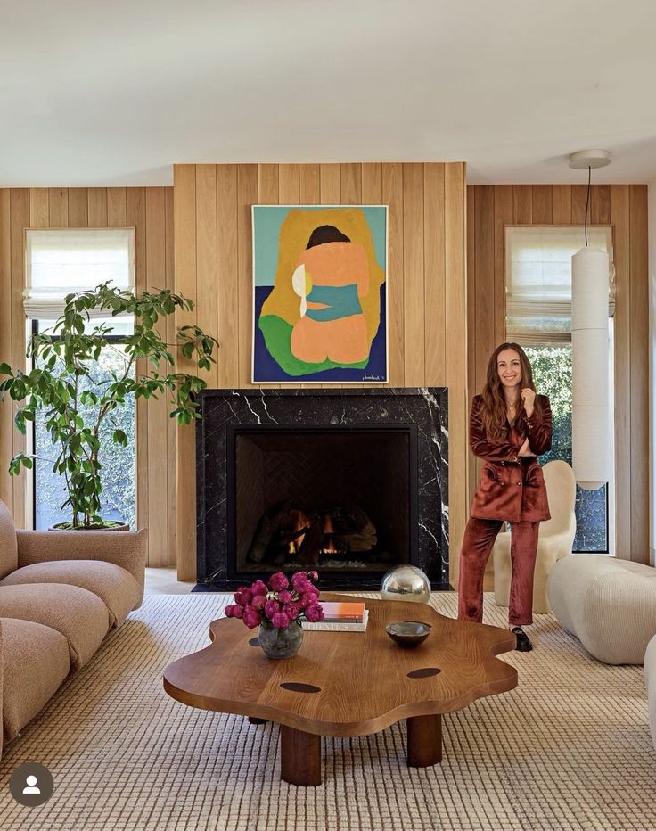 a woman standing in a living room next to a fire place with a painting on the wall