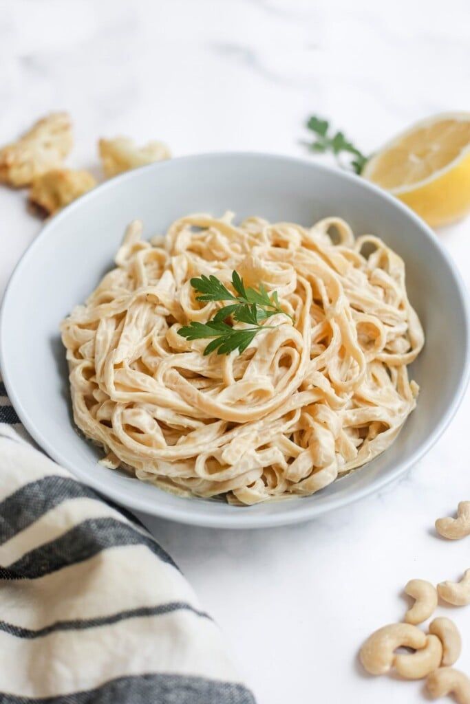 a white bowl filled with pasta and garnished with parsley next to cashews