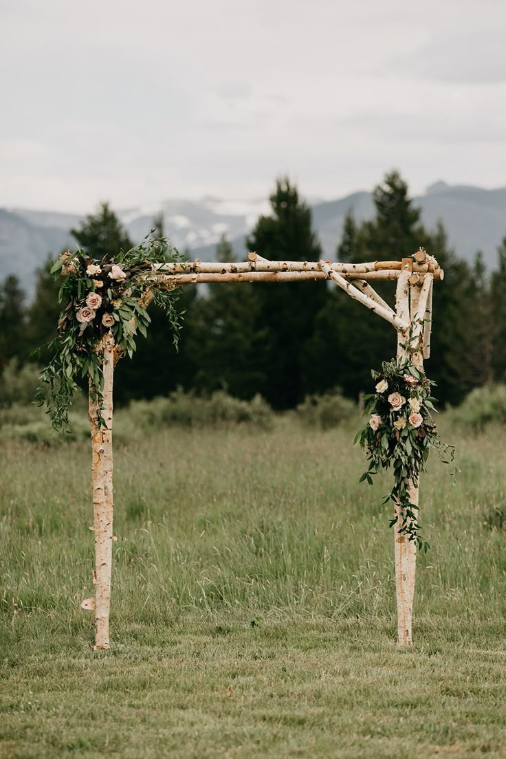 a wooden arch with flowers and greenery on the grass in front of some mountains