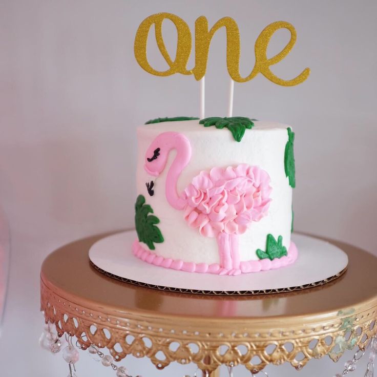 a pink flamingo cake with one candle on top and the number one in gold