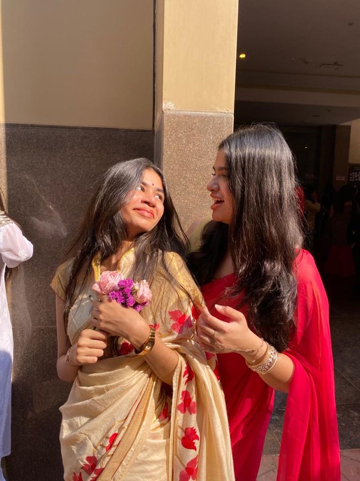 two women standing next to each other in front of a building and one is holding a bouquet of flowers