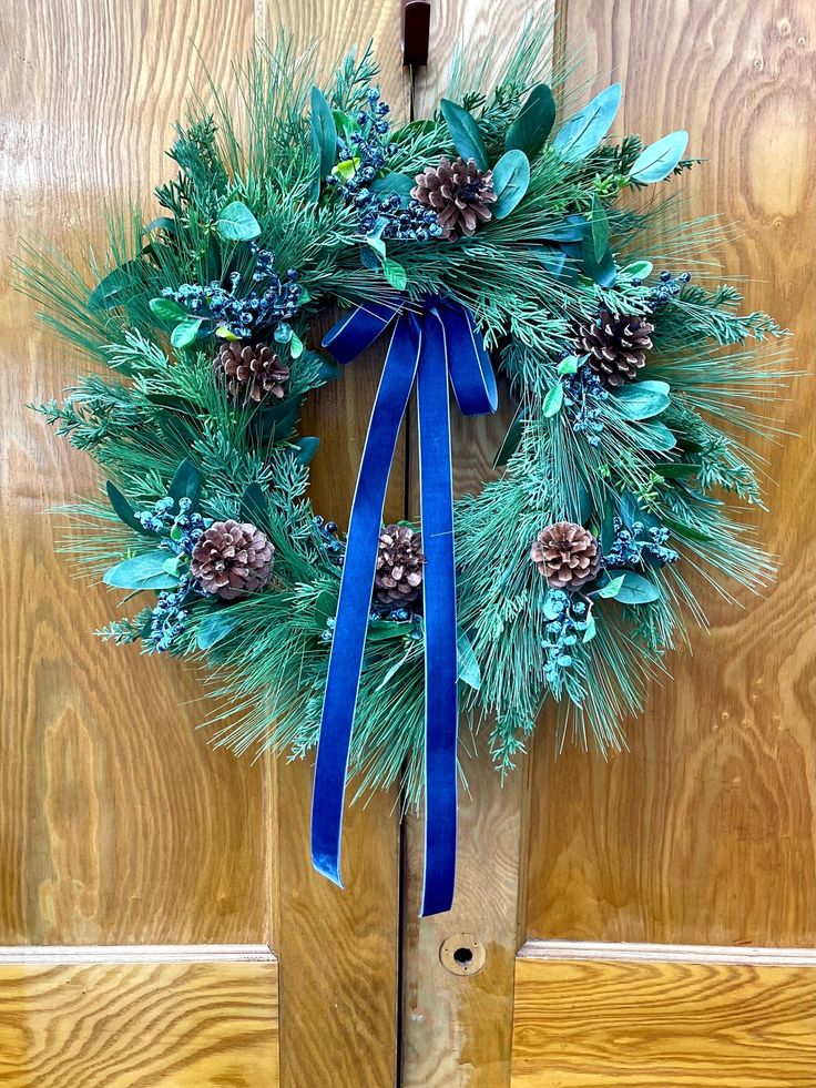 a wreath with pine cones and blue ribbon hanging on a door