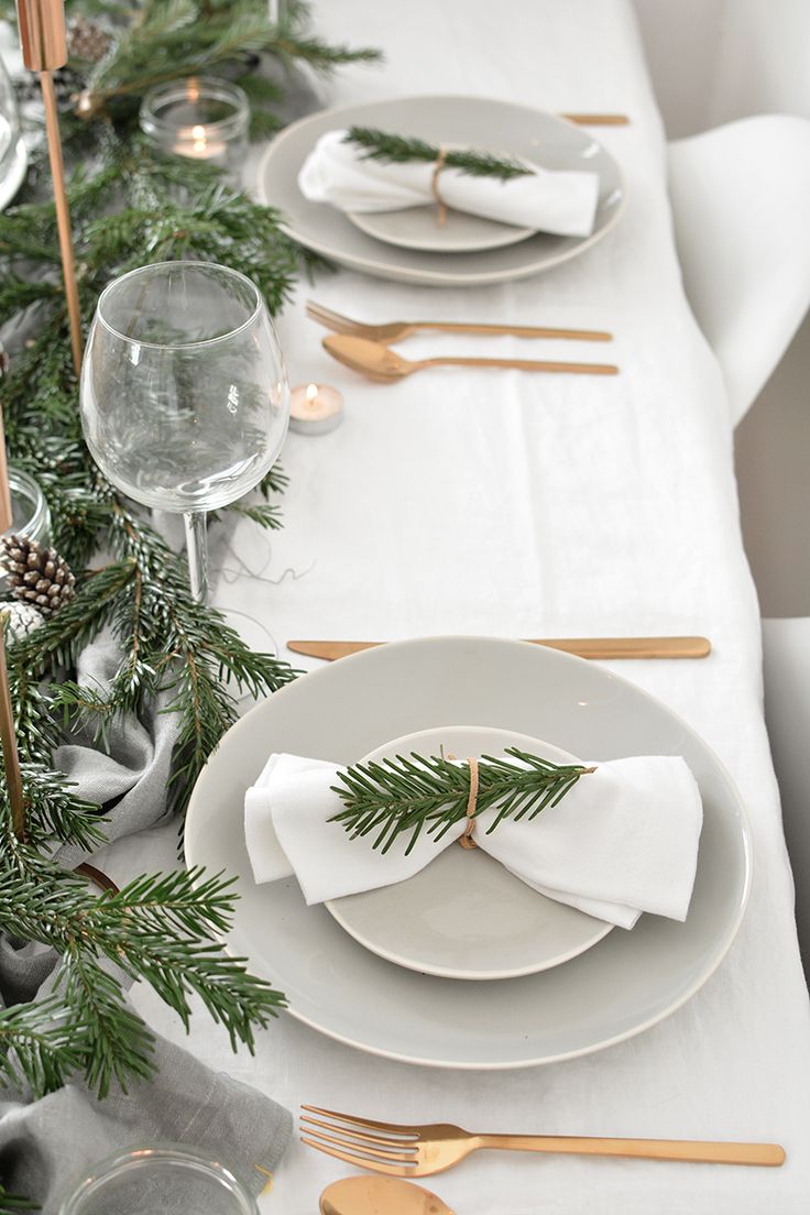 the table is set with white plates, silverware and pine branches on it for christmas dinner