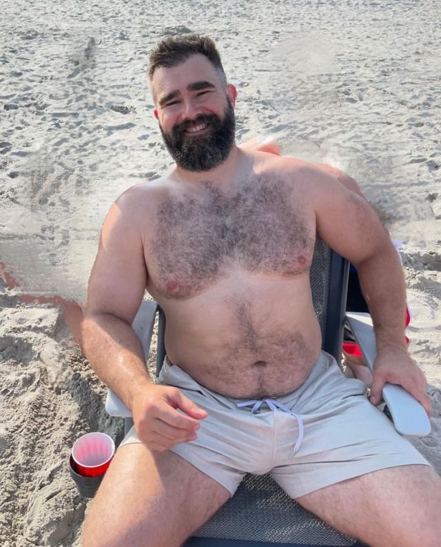 a shirtless man sitting in a chair on the beach with his cup and drink