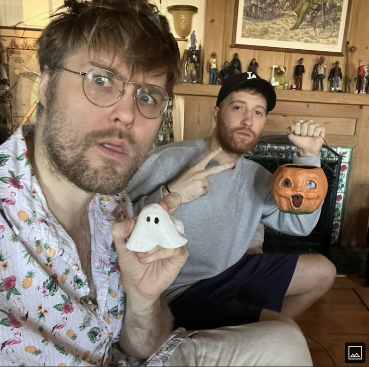 two men sitting on a couch with fake ghost heads in front of them and one holding a pumpkin