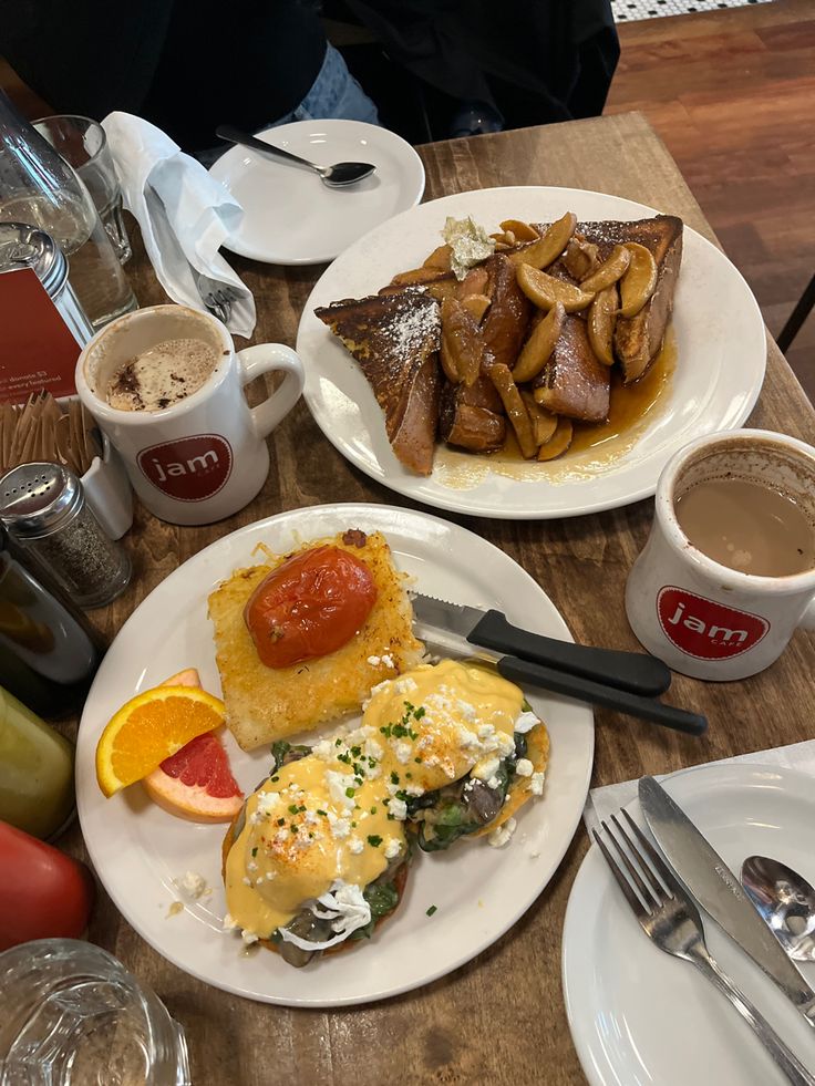 two white plates topped with food next to cups of coffee and saucers on a wooden table