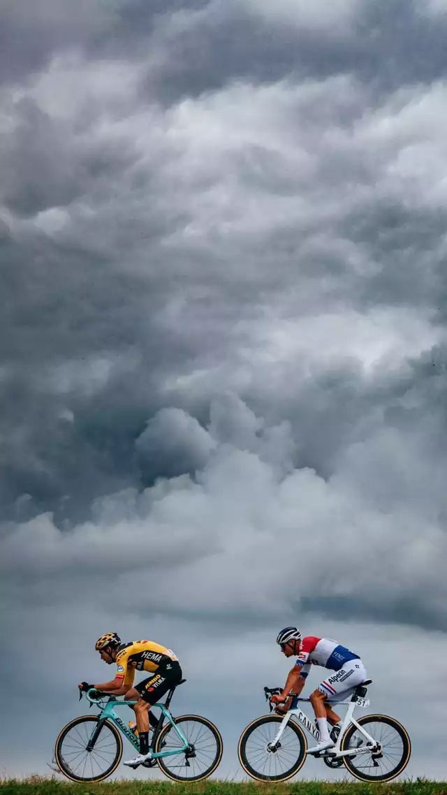 two bicyclists are riding in the grass under a cloudy sky with dark clouds