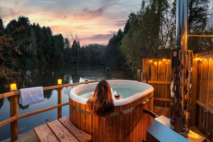 a woman sitting in a wooden hot tub next to a body of water at sunset