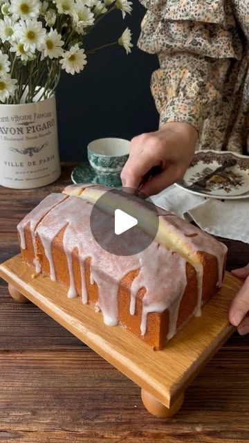 a person cutting a loaf of cake on top of a wooden table with flowers in the background