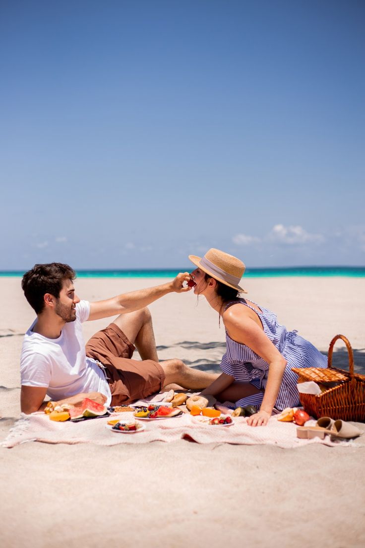 a man and woman sitting on the beach with food in front of them, eating