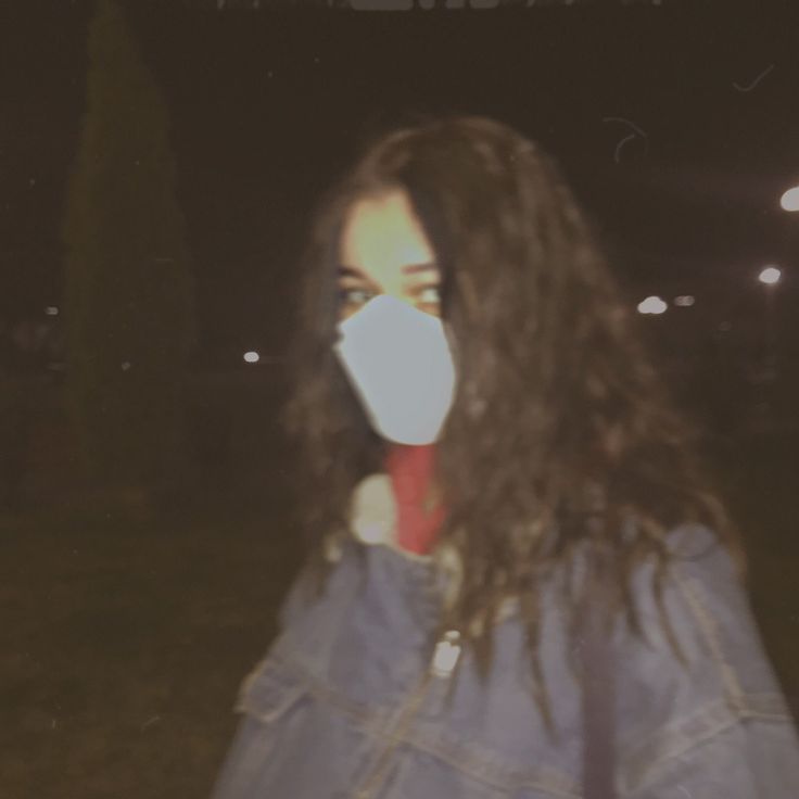 a woman with long dark hair wearing a white face mask and blue denim jacket is standing in the grass at night