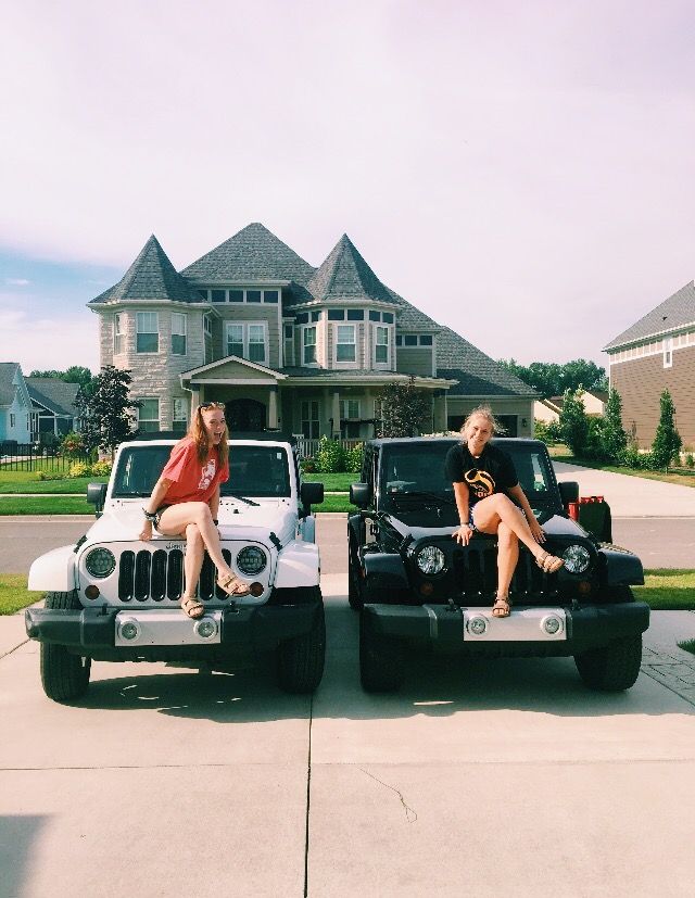 two women sitting on the hood of their jeeps in front of a large house