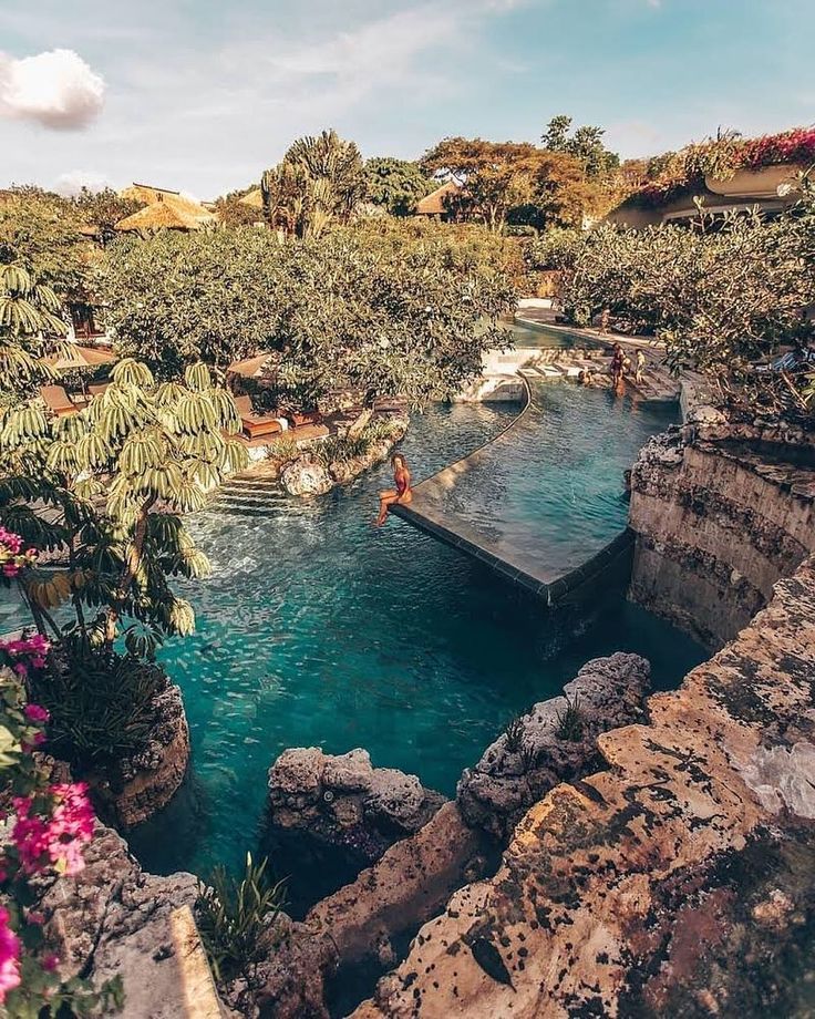 a man is swimming in a pool surrounded by trees and flowers on the side of a cliff