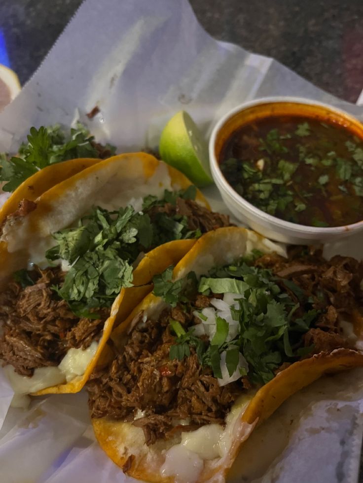 three tacos with meat, cheese and cilantro on paper next to a bowl of salsa