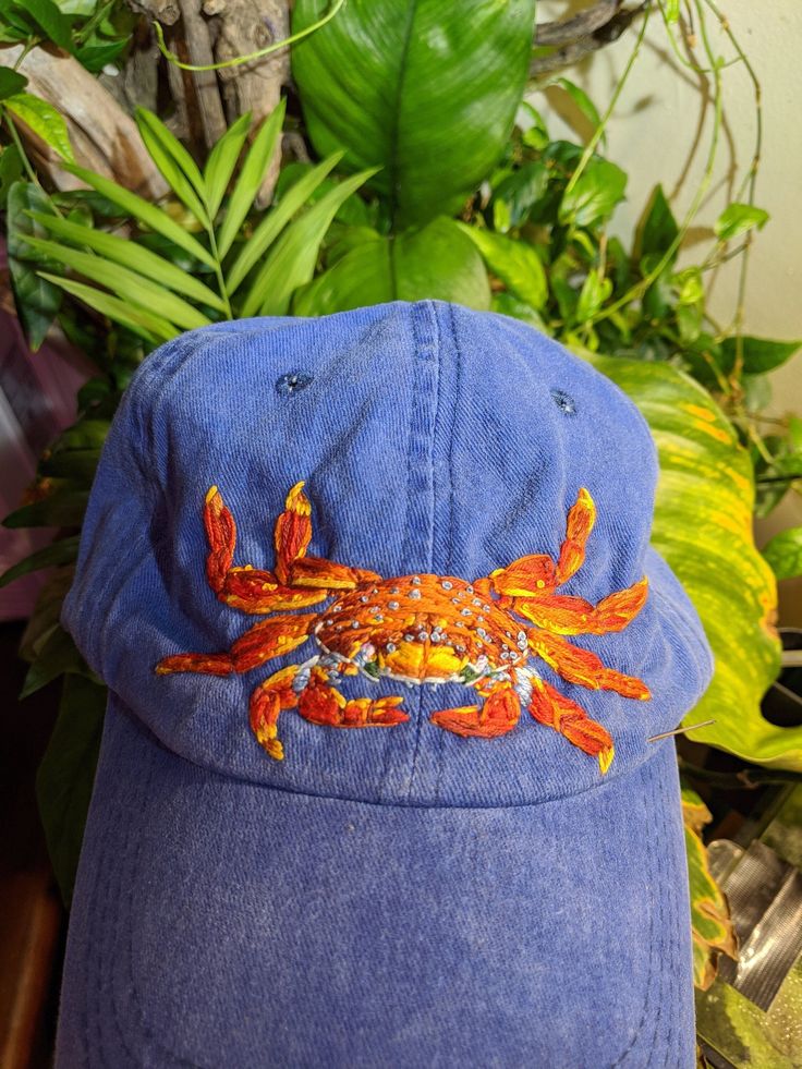 a blue hat with an orange crab embroidered on the front and side, sitting next to green plants