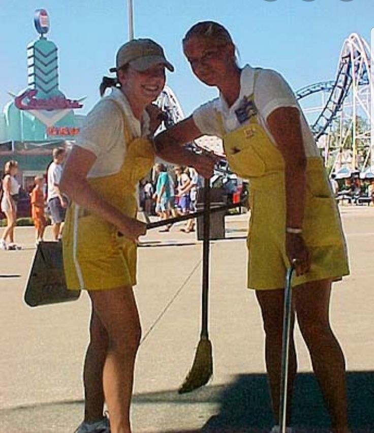 two people dressed in yellow posing for a photo at an amusement park, one holding a broom and the other smiling