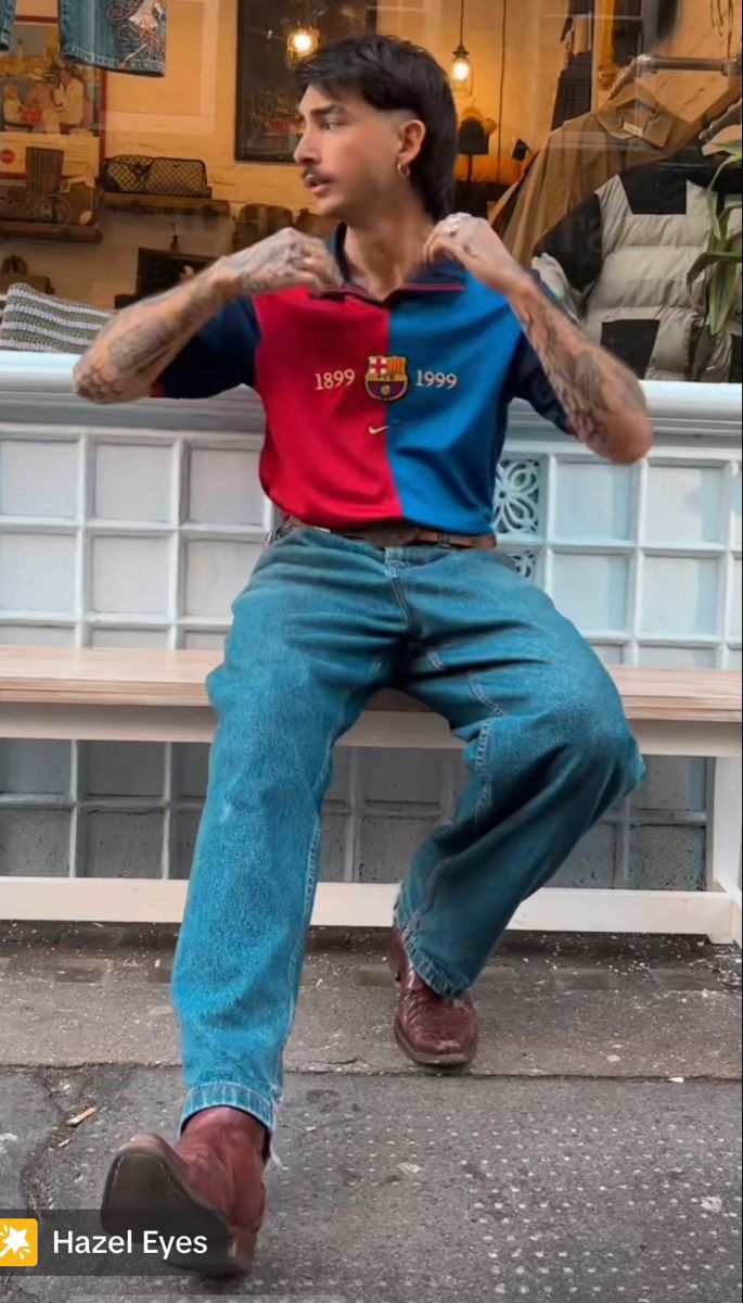 a man with tattoos sitting on a bench in front of a store window wearing blue jeans and a red shirt