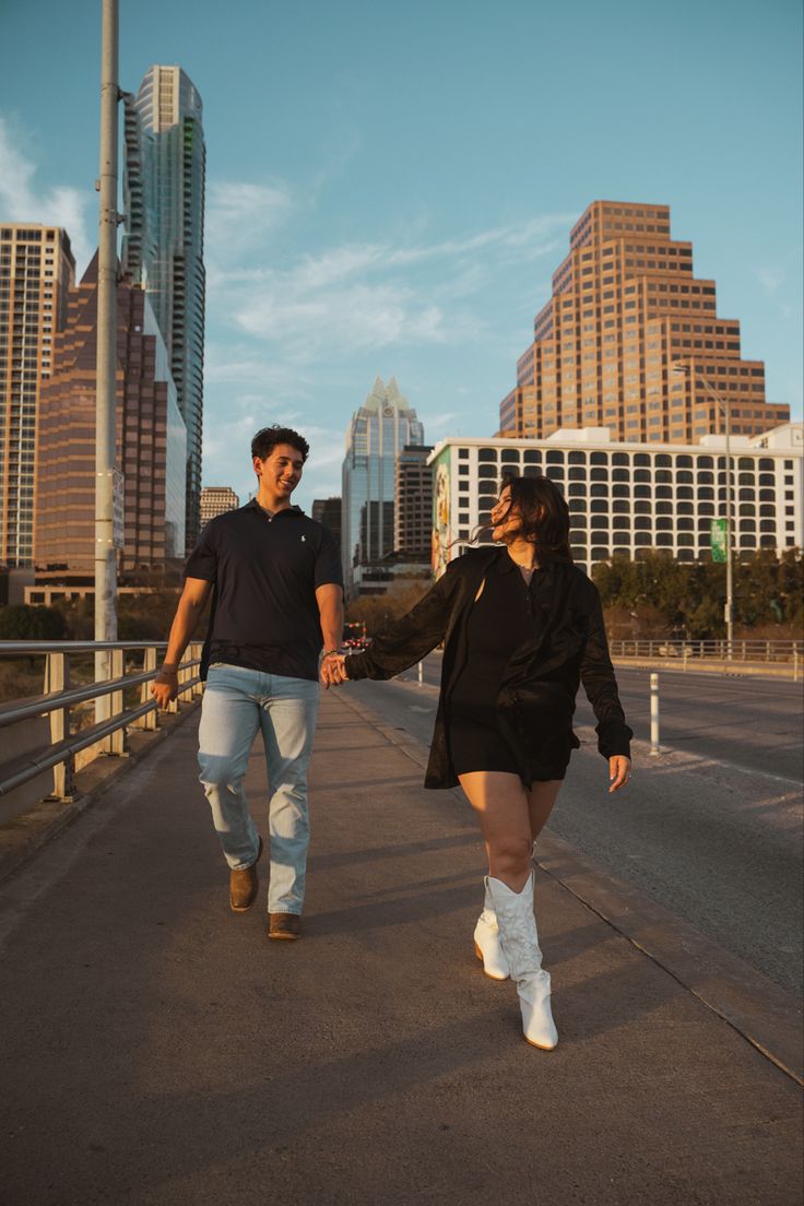 a man and woman holding hands walking down the street with tall buildings in the background