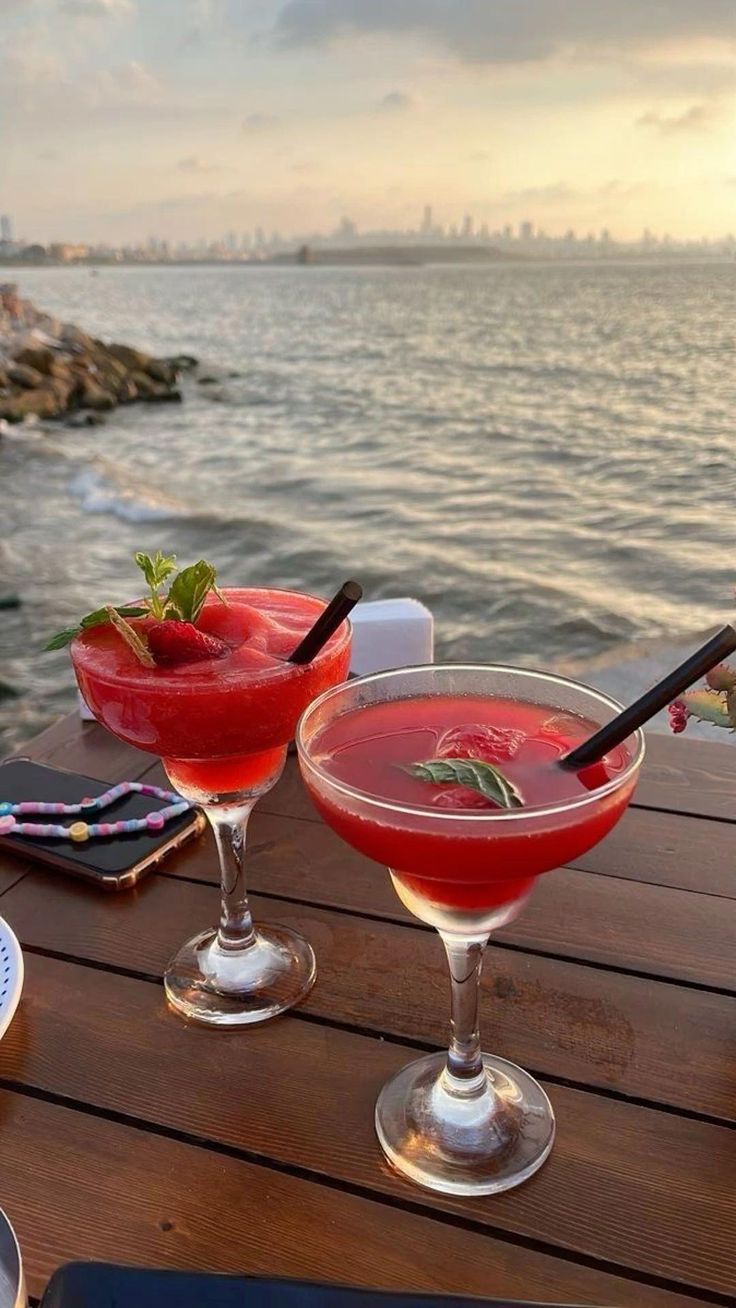 two cocktails sitting on top of a wooden table next to the ocean