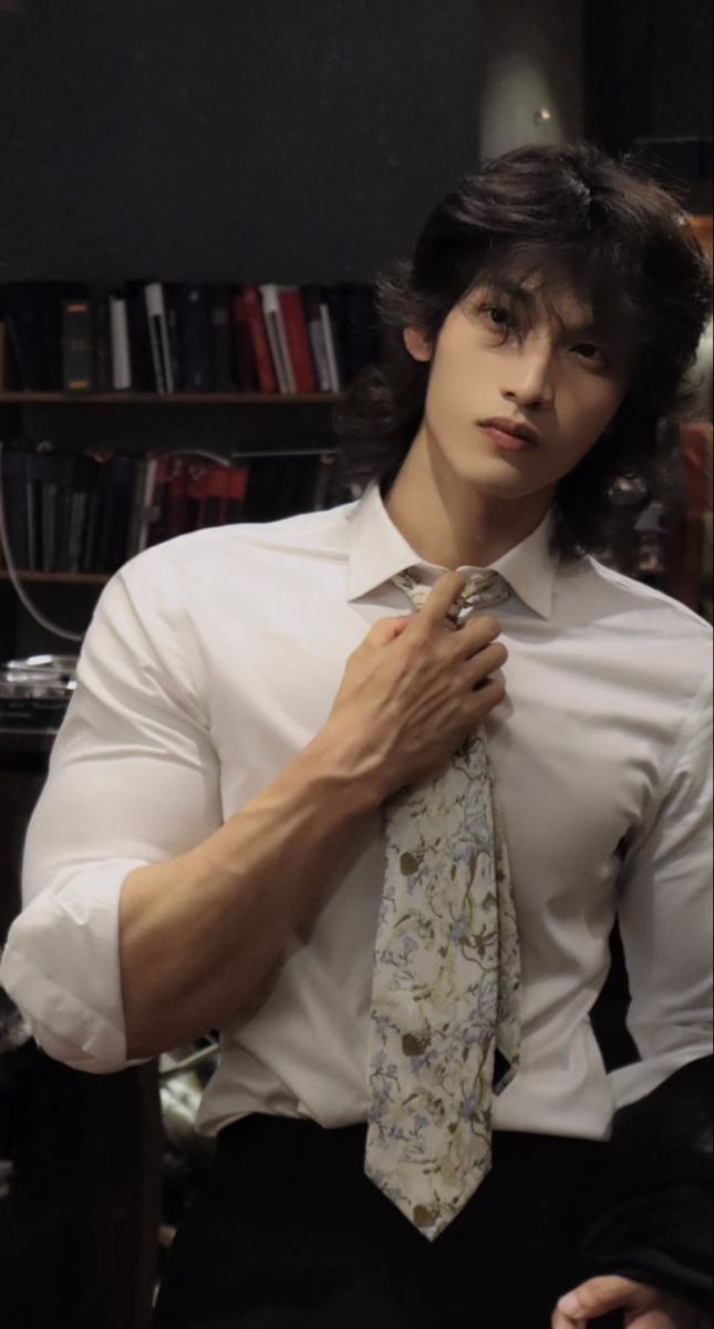 a young man wearing a white shirt and tie with his hands on his chest, in front of a bookshelf