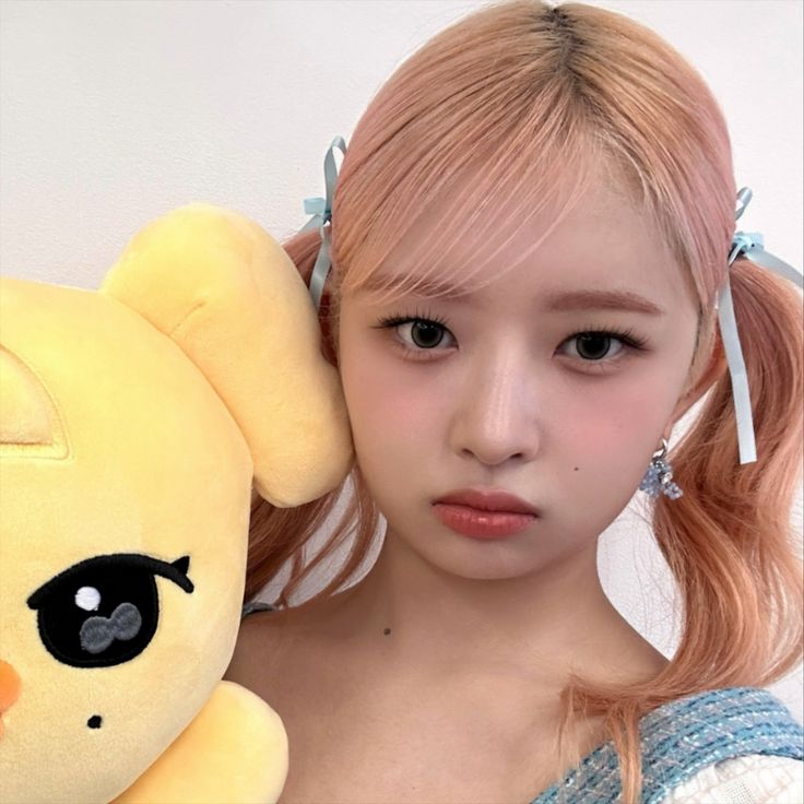 a girl with pink hair holding a yellow stuffed animal in front of her face and looking at the camera