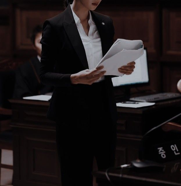 a woman standing in front of a desk holding papers and looking at a computer screen