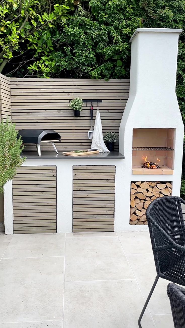 an outdoor fireplace and grill in the middle of a patio area with chairs, table and firewood logs