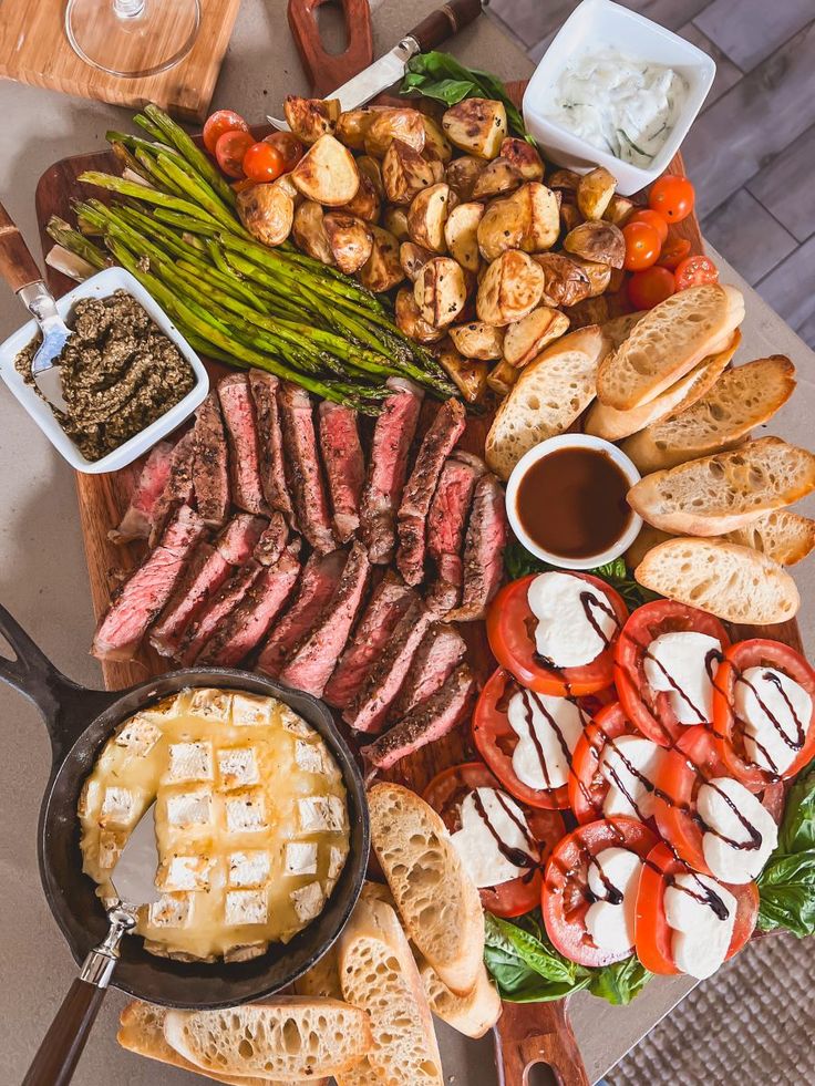 an assortment of meats, vegetables and bread on a wooden platter with utensils