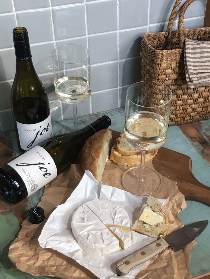 two glasses of wine, cheese and bread on a wooden tray next to bottles of wine