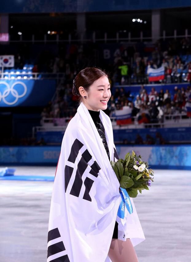 a woman is standing on the ice with flowers in her hand and an olympic flag draped over her shoulders