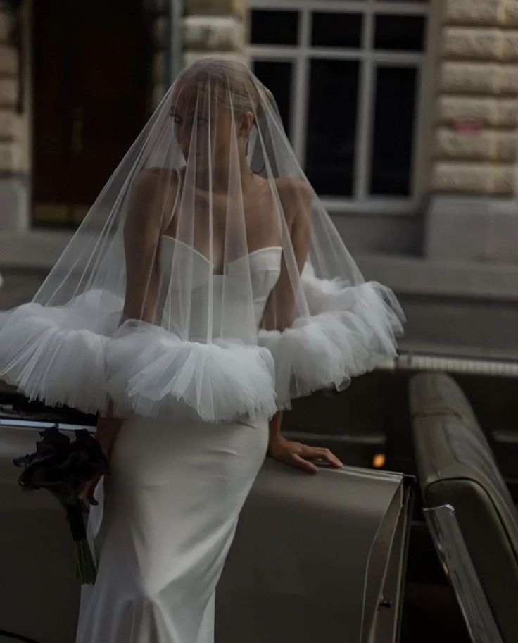 a woman in a white wedding dress and veil leaning on a car with her back to the camera