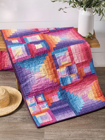 a colorful quilt and hat sitting on a wooden floor next to a vase filled with flowers