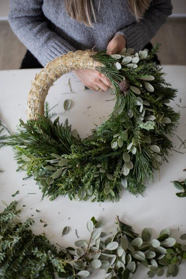 a woman is making a wreath with greenery and pine cones on a white table