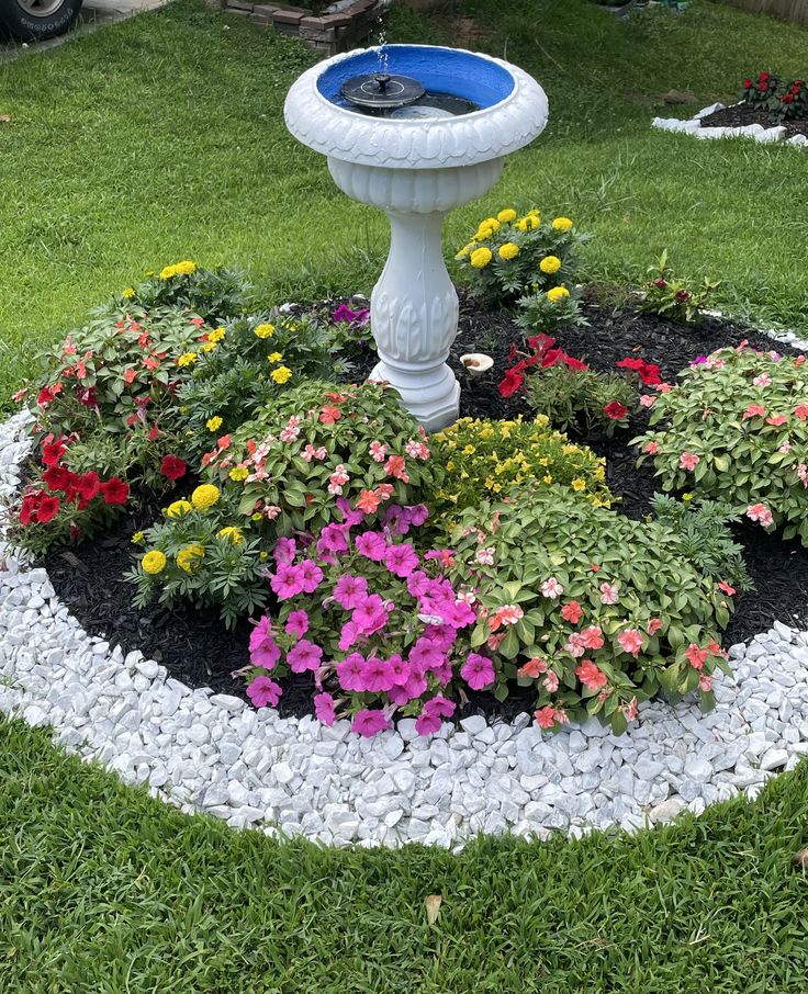 a bird bath in the middle of a flower bed with flowers around it and rocks