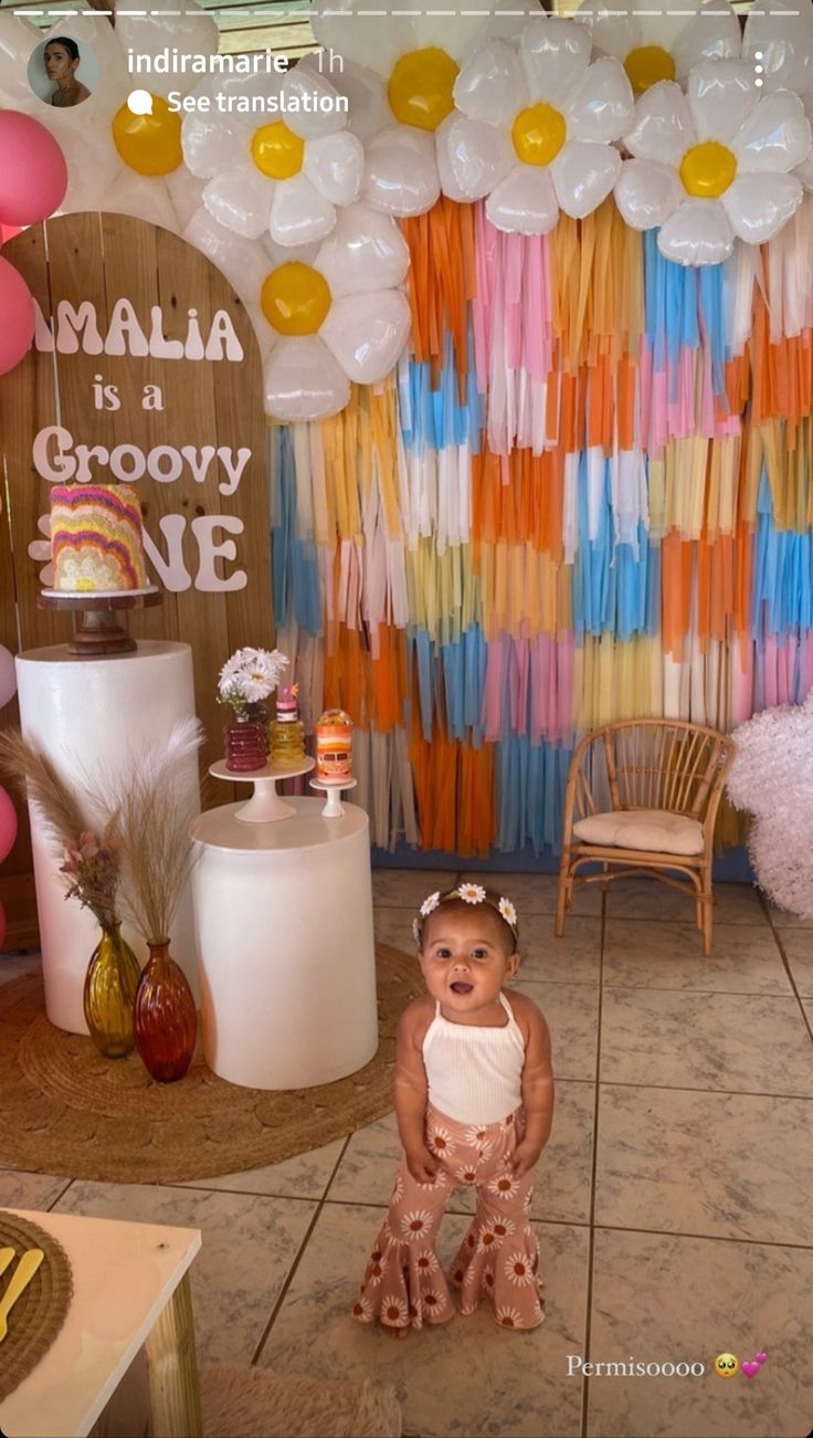 a baby girl standing in front of a cake and balloons display at a birthday party