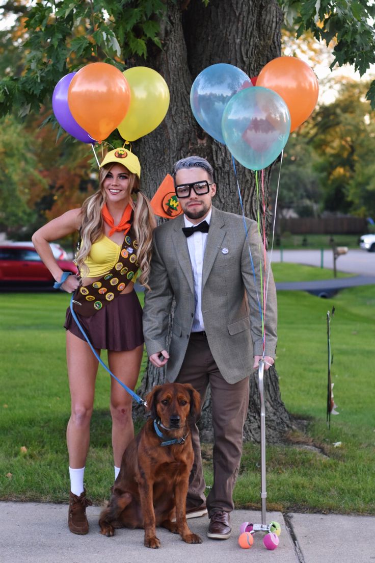 a man and woman standing next to a dog with balloons on their heads, in front of a tree