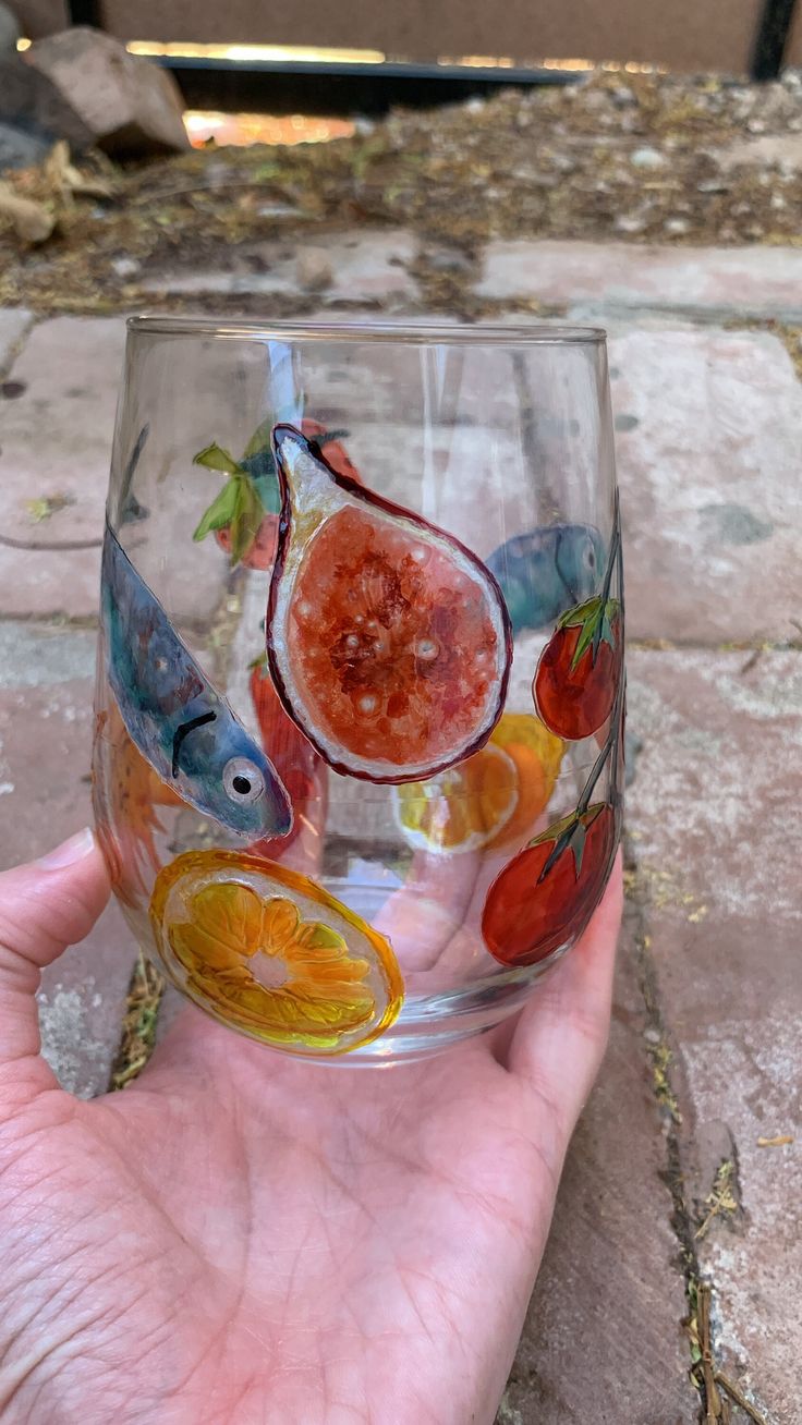 a hand holding a wine glass with fruit painted on the side and fish in it