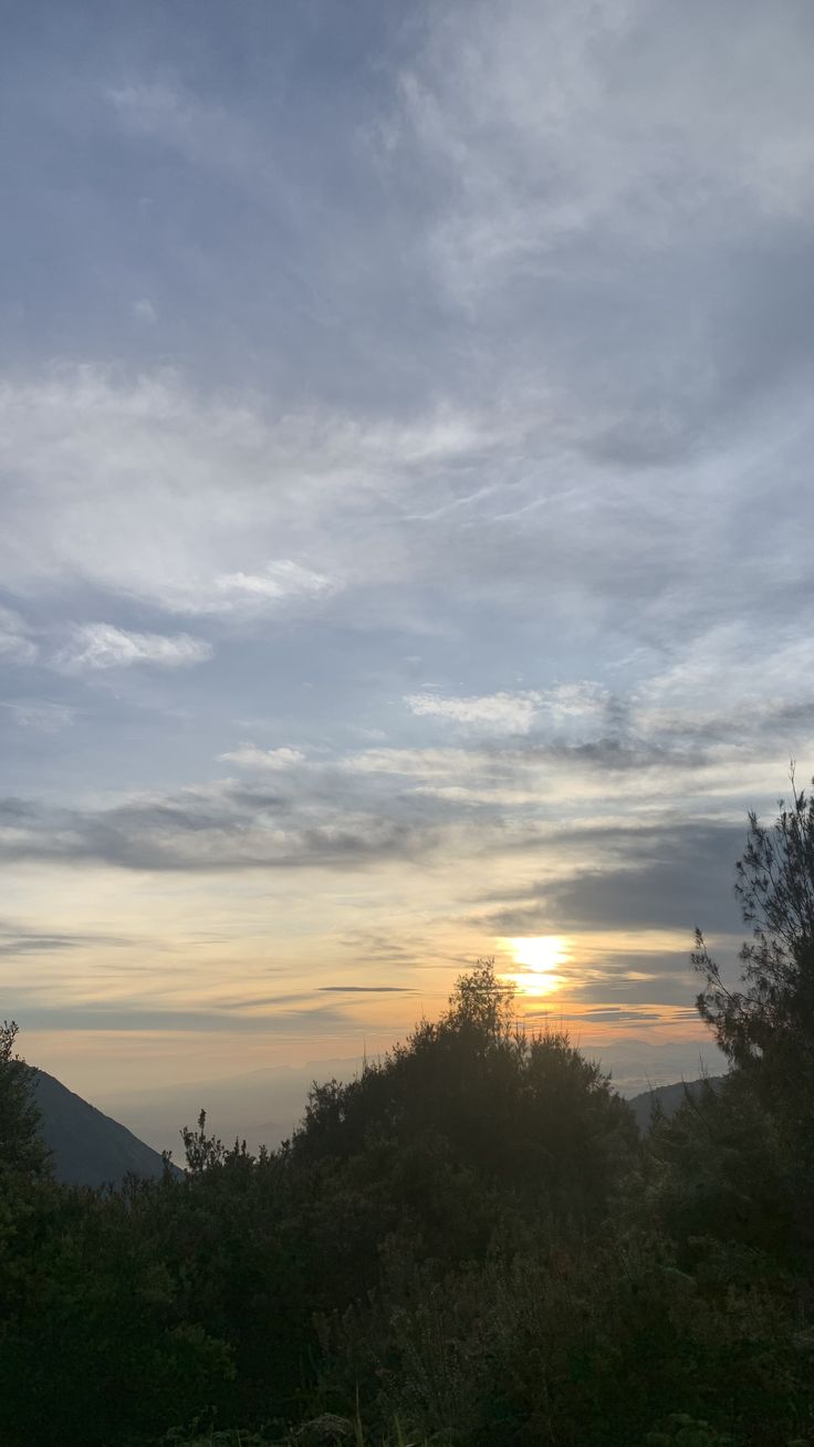 the sun is setting over some trees and hills in the distance, with mountains in the background