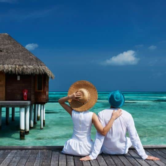 two people sitting on a dock looking out at the ocean with huts in the background