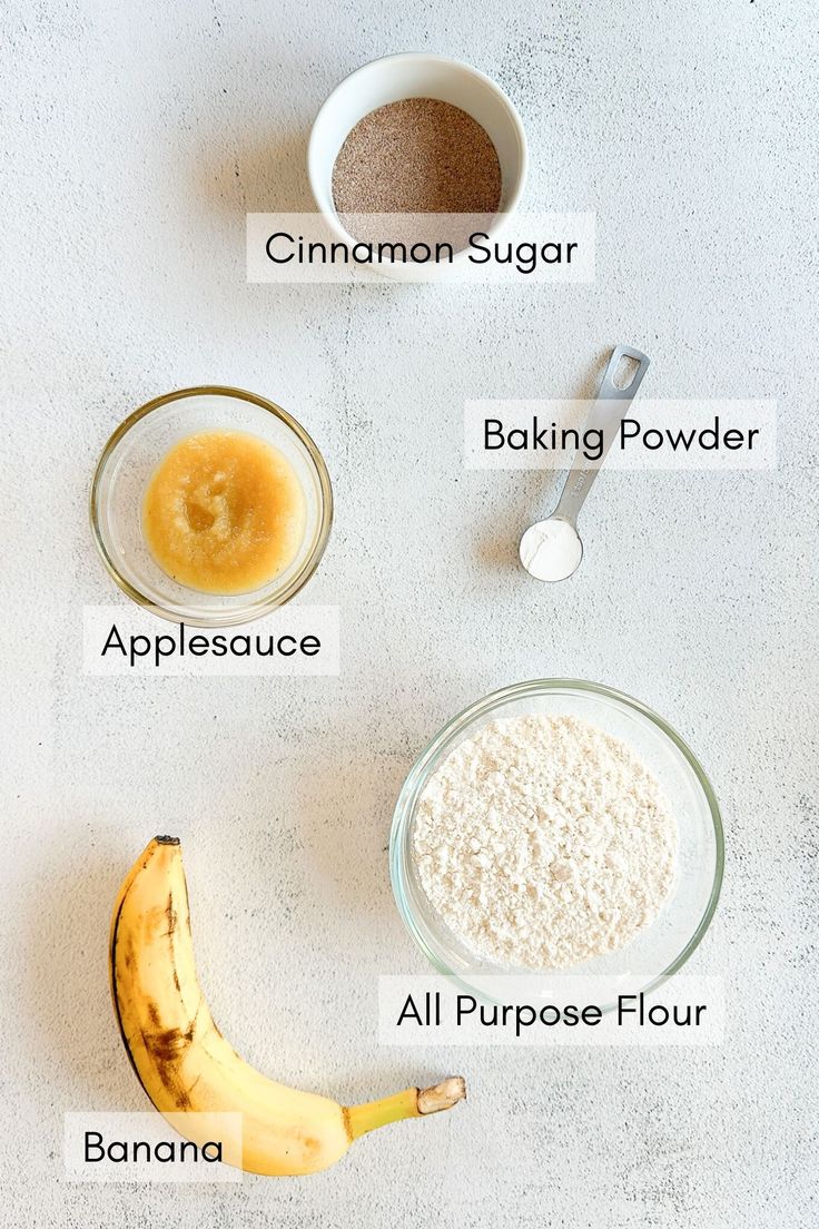 ingredients to make banana bread laid out on a white counter top, including bananas, flour, sugar, baking powder, and an all purpose flour