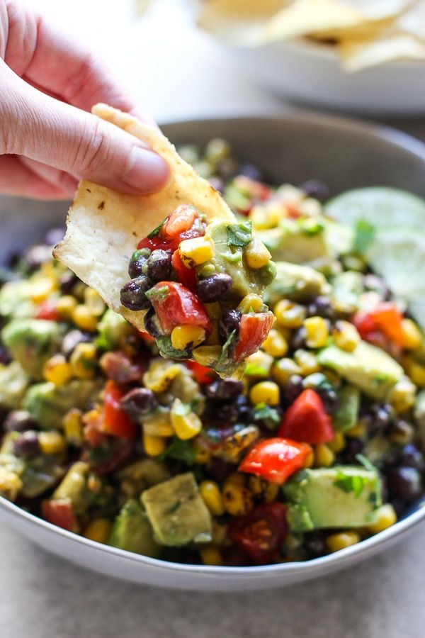 a hand holding a tortilla chip over a bowl of black beans, corn and avocado salsa