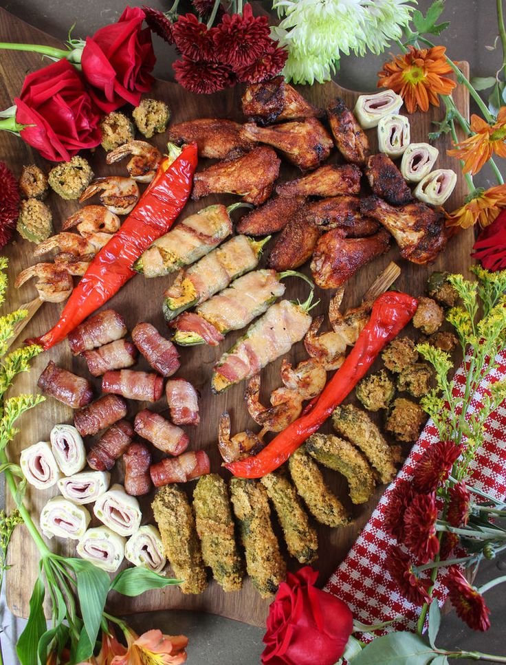 an assortment of meats and vegetables on a wooden cutting board with flowers in the background