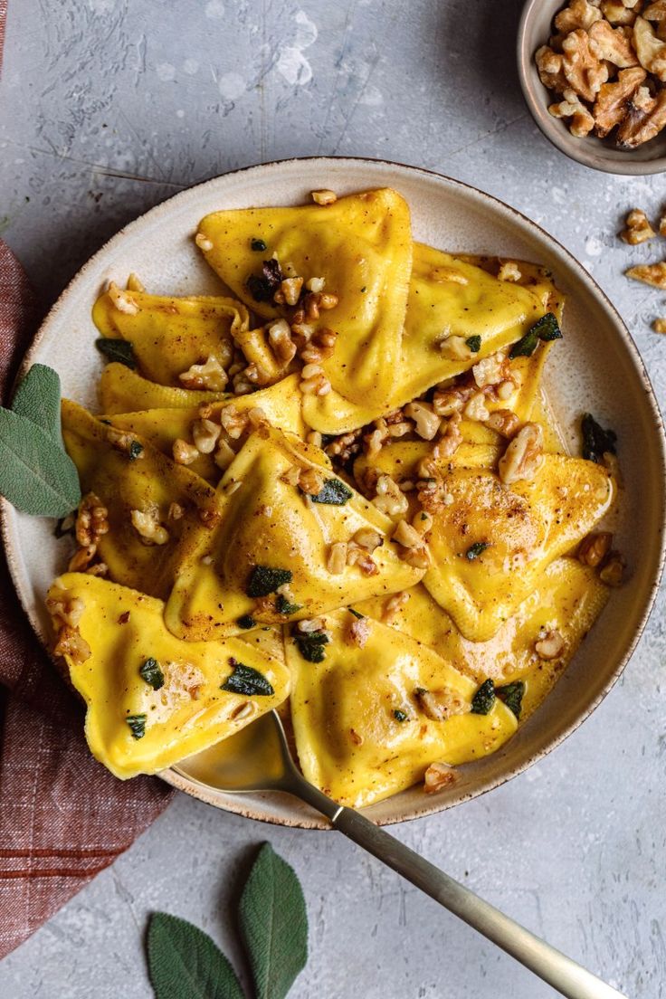 a white plate topped with ravioli and walnuts next to a bowl of nuts