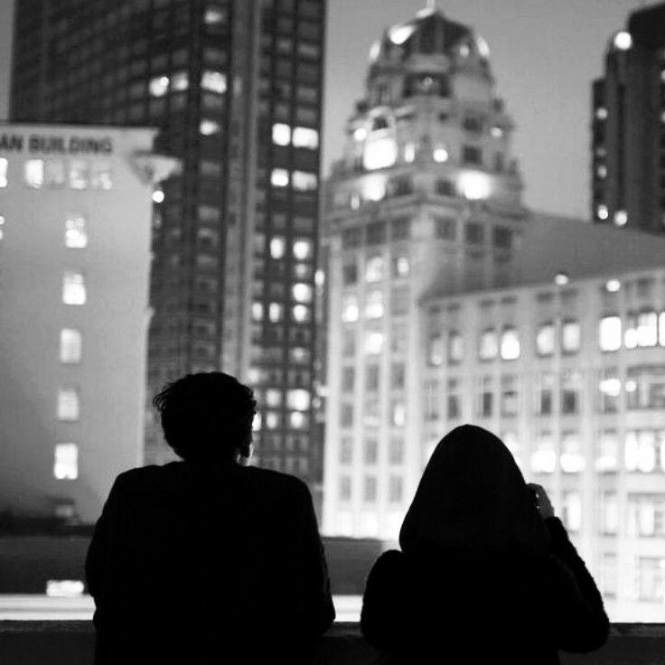 two people are sitting on a ledge looking at the city lights in black and white