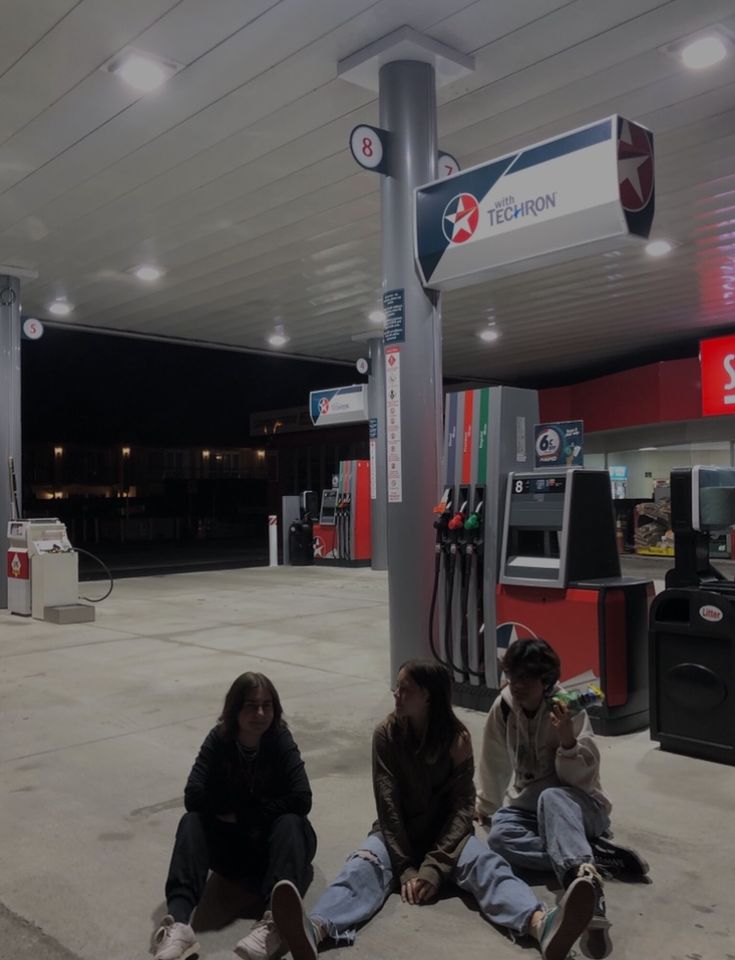 three people sitting on the ground in front of a gas station