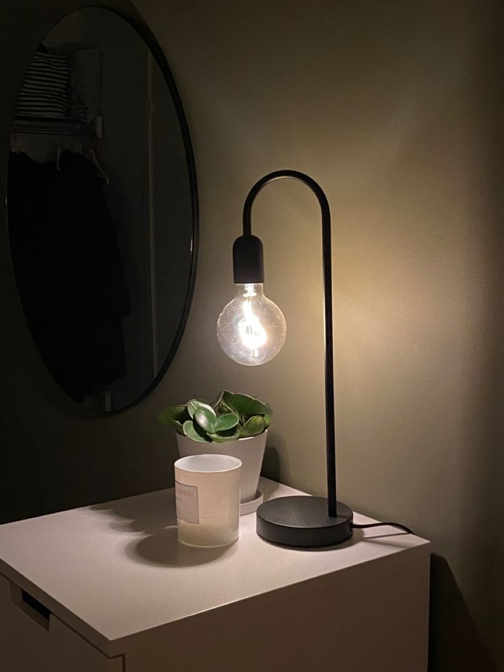 a light that is on top of a white dresser next to a mirror and potted plant