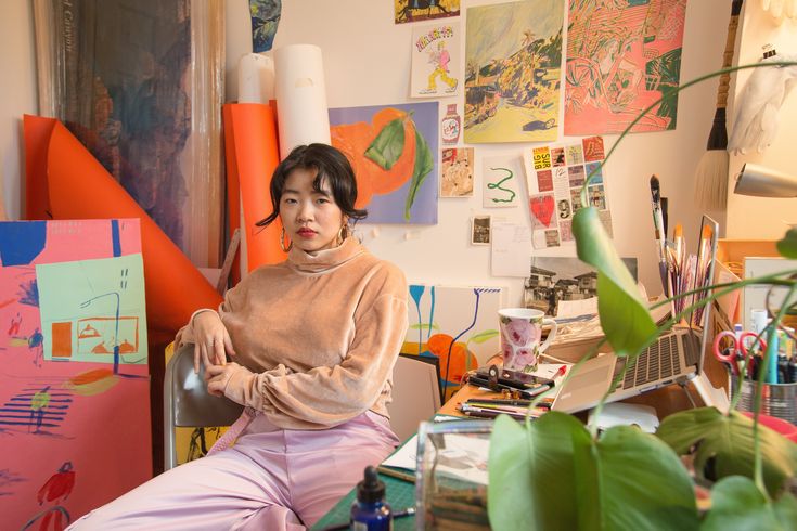 a woman sitting on a chair in front of a desk covered with art and plants