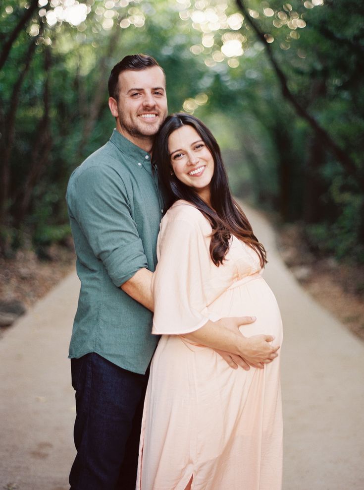 a pregnant woman and man pose for a photo in front of trees on a path